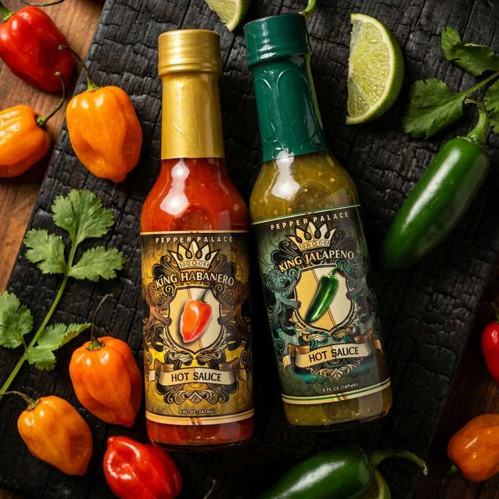 Two bottles of Pepper Palace hot sauce, King Habanero and King Jalapeño, arranged on a dark textured surface with fresh peppers, cilantro, and lime wedges.